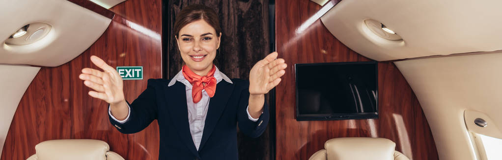 panoramic shot of smiling flight attendant in uniform with outstretched hands in private plane  - 写真・画像
