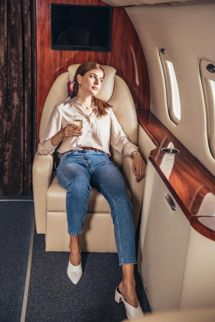 aantrekkelijke vrouw in shirt met champagne glas en kijken door het raam in prive-vliegtuig  - Foto, afbeelding