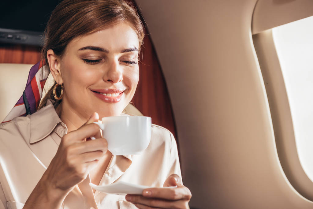 sonriente mujer en camisa sosteniendo taza de café en avión privado
  - Foto, Imagen