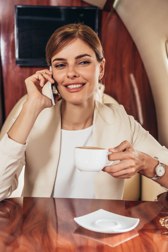 aantrekkelijke zakenvrouw in pak holding cup en praten op de smartphone in prive-vliegtuig  - Foto, afbeelding
