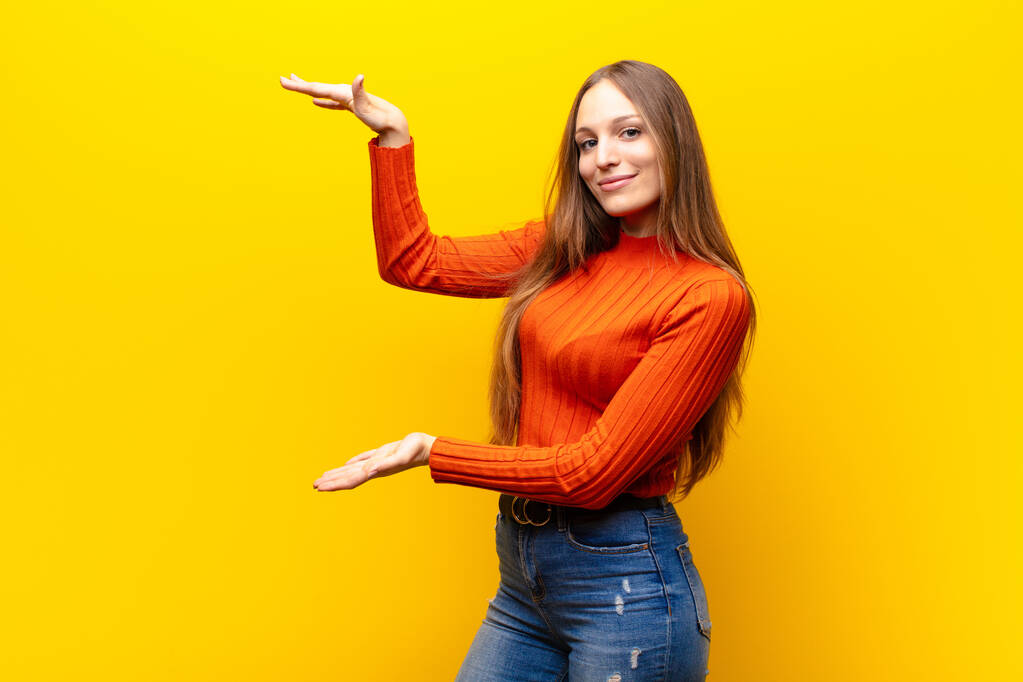 young pretty woman smiling, feeling happy, positive and satisfied, holding or showing object or concept on copy space against orange background - 写真・画像
