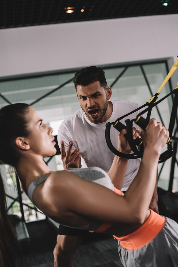 excited trainer shouting while motivating young sportswoman pulling up on suspension trainer - Photo, Image