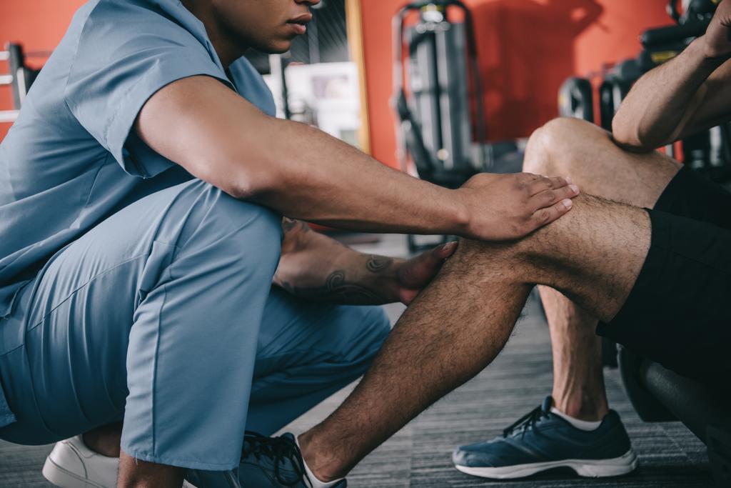bijgesneden beeld van Afrikaans-Amerikaanse arts raken gewond knie van sportman  - Foto, afbeelding