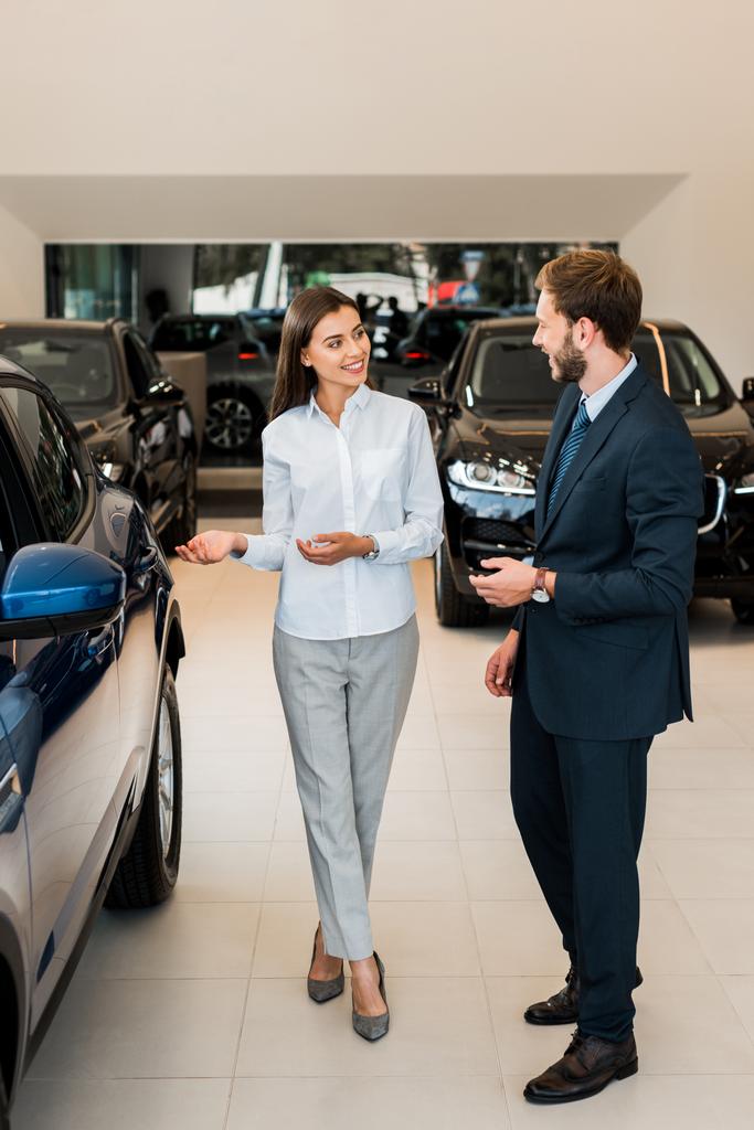 cheerful car dealer gesturing while looking at bearded man in car showroom  - Photo, Image