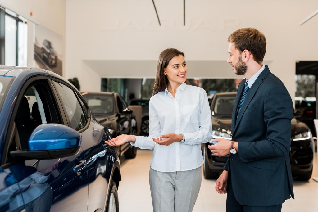 positive car dealer gesturing while looking at bearded man in car showroom  - Photo, Image