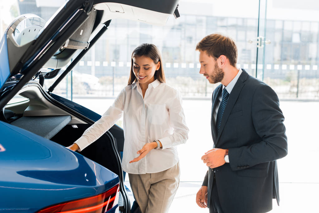 attractive and happy car dealer gesturing near blue car and bearded man  - Photo, Image