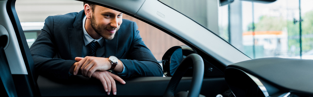panoramiczne ujęcie szczęśliwego brodatego mężczyzny patrzącego na nowoczesny samochód  - Zdjęcie, obraz