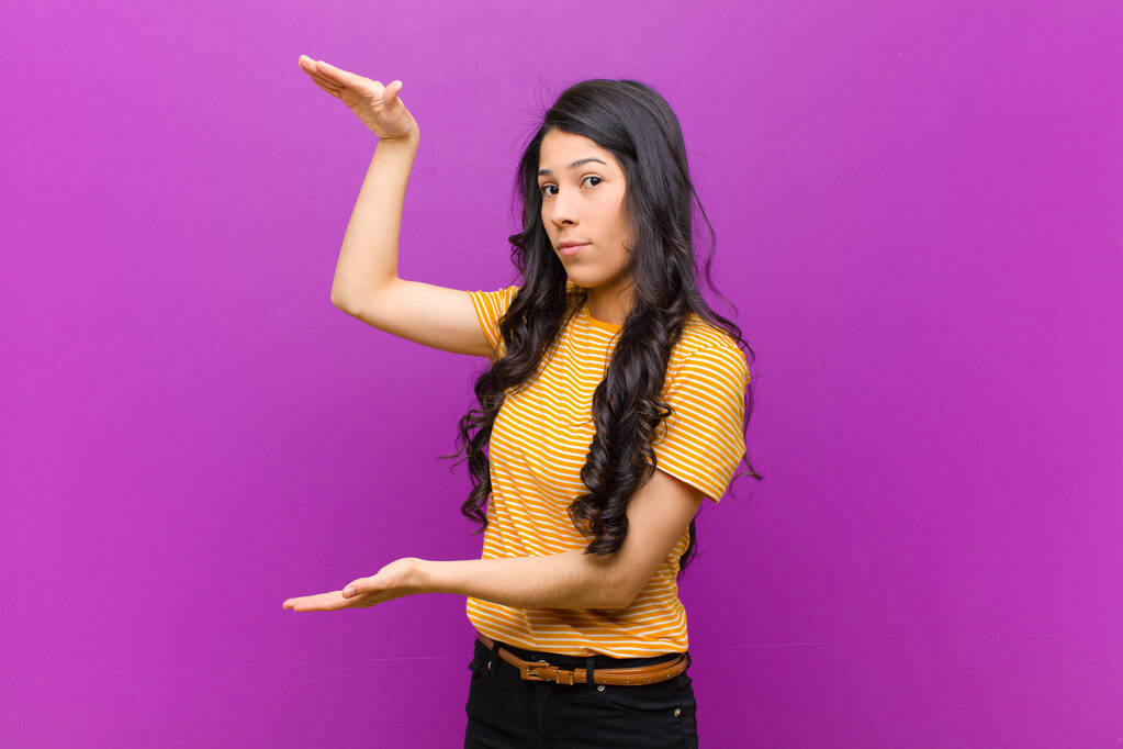 mladá hezká latinská žena drží objekt s oběma rukama na straně kopírovat prostor, ukazuje, nabízí nebo propaguje objekt proti fialové zdi - Fotografie, Obrázek