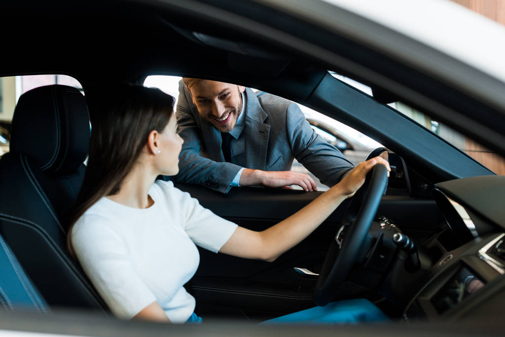 selectieve focus van lachende vrouw zittend in auto in de buurt van bebaarde man in de auto showroom  - Foto, afbeelding