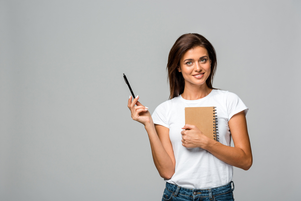 Lächelnde junge Frau mit Stift und Notizbuch, isoliert auf grau - Foto, Bild