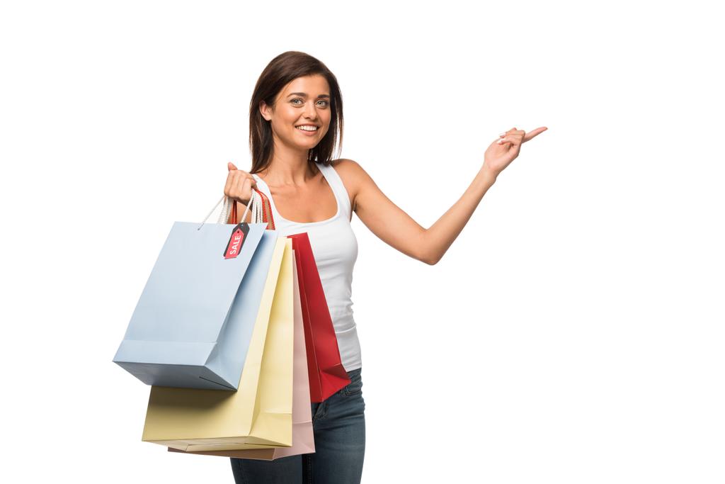 smiling girl holding shopping bags with sale signs while pointing isolated on white  - 写真・画像