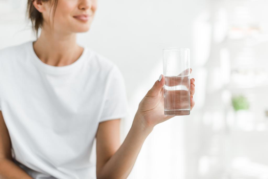 καλλιεργημένη άποψη του χαμογελαστού κοριτσιού κρατώντας ένα ποτήρι νερό το πρωί - Φωτογραφία, εικόνα