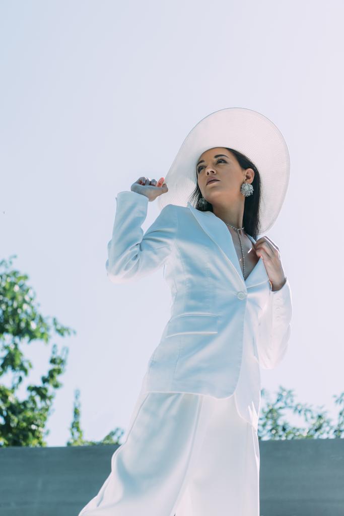 visão de ângulo baixo de mulher atraente em terno branco e chapéu posando fora
  - Foto, Imagem