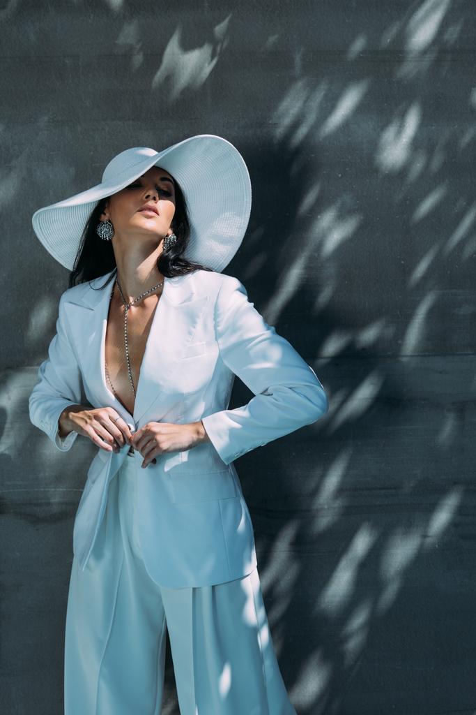 attraktive Frau in weißem Anzug und Hut posiert mit geschlossenen Augen draußen  - Foto, Bild