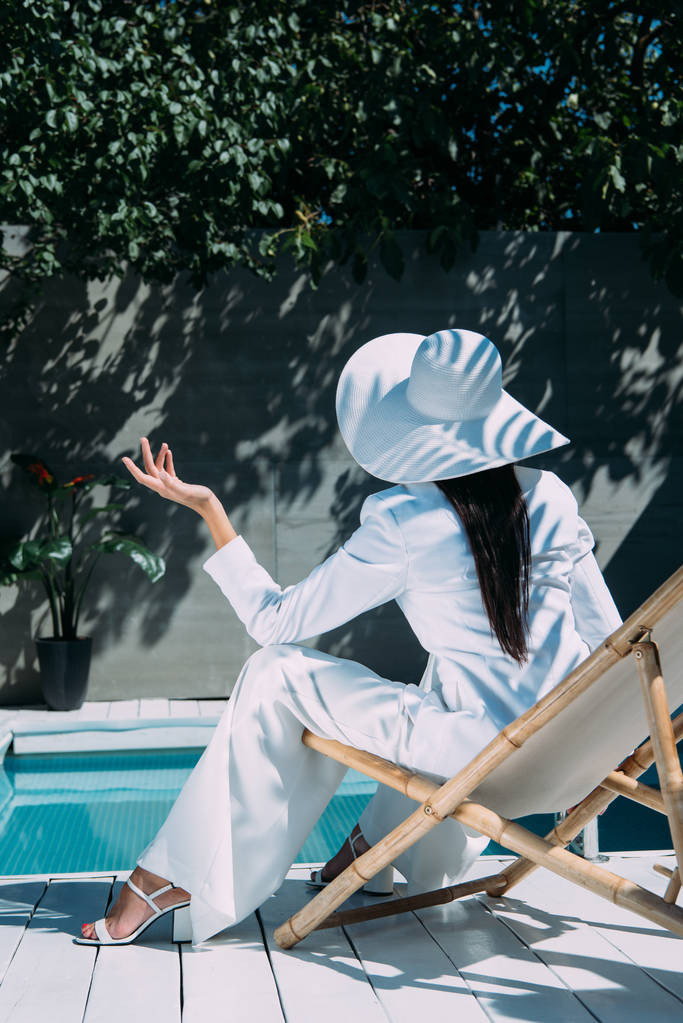 вид ззаду жінки в білому костюмі і капелюсі, що сидить на стільці зовні
  - Фото, зображення