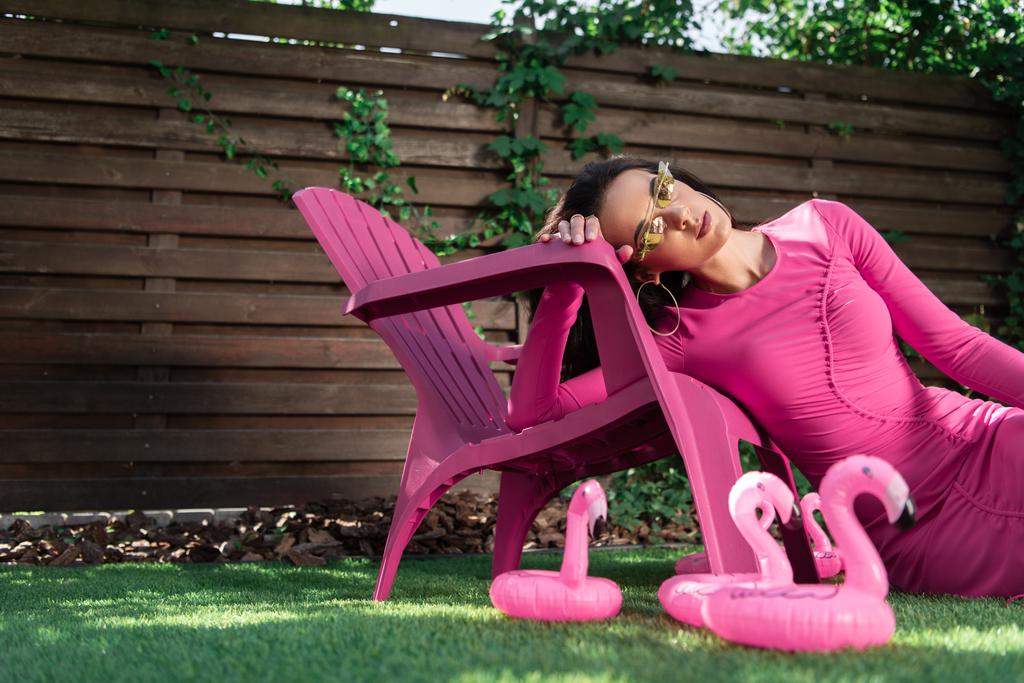 atraktivní žena v šatech a slunečních brýlích pózuje a sedí na trávě venku  - Fotografie, Obrázek