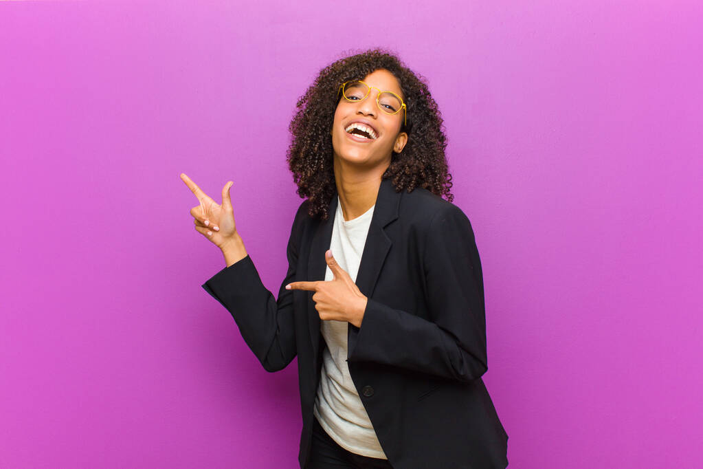 junge schwarze Geschäftsfrau, die freudig und überrascht ist, mit schockiertem Gesichtsausdruck lächelt und zur Seite zeigt - Foto, Bild