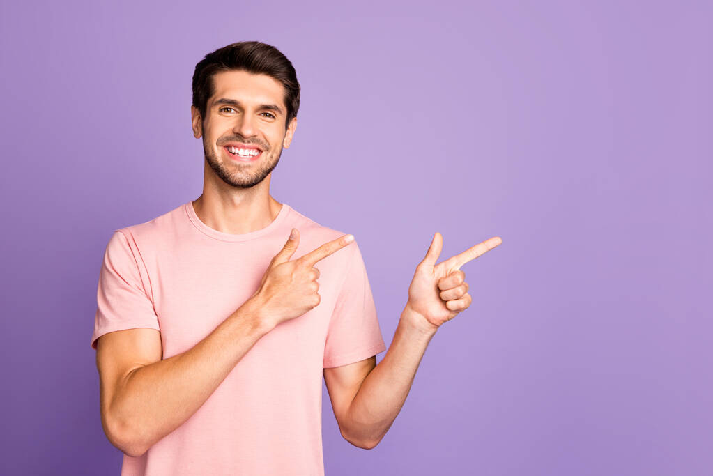 Portrét jeho hezký atraktivní veselý sebevědomý vousatý chlap na sobě růžové tričko ukazující dvěma ukazováčky stranou, jako sledovat předplatné izolované na fialové fialové fialové pastelové barvy pozadí - Fotografie, Obrázek
