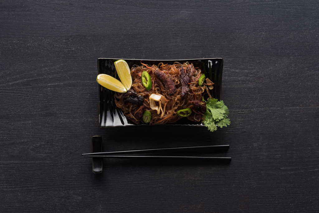 bovenaanzicht van smakelijk kruidig vlees thai noedels bij eetstokjes op houten grijze ondergrond - Foto, afbeelding