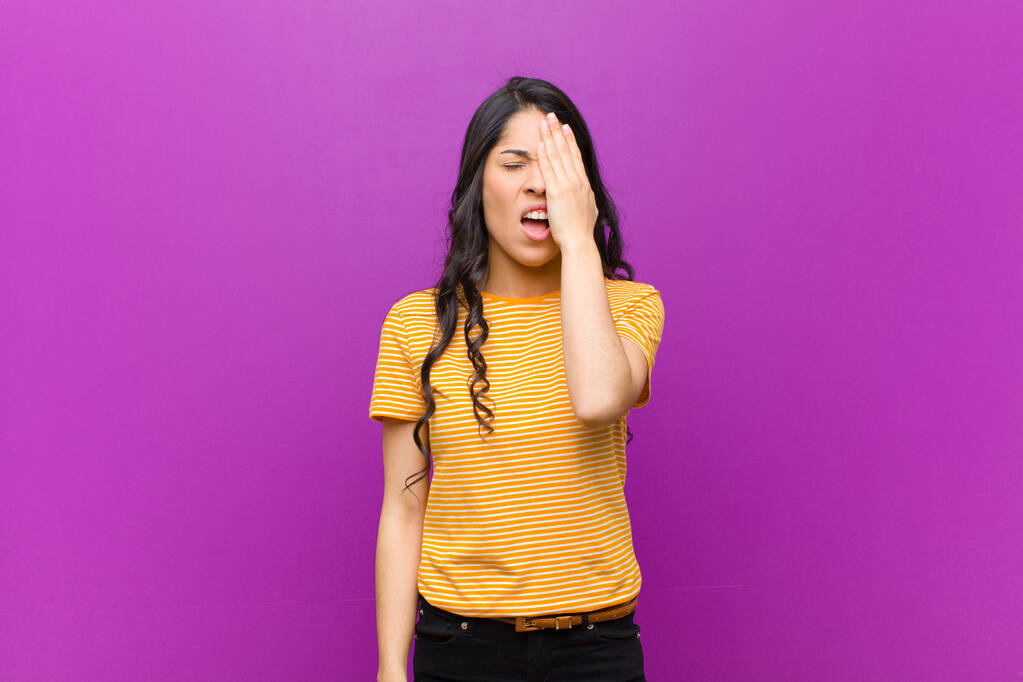 junge hübsche lateinamerikanische Frau sieht schläfrig, gelangweilt und gähnend aus, mit Kopfschmerzen und einer Hand, die das halbe Gesicht gegen eine violette Wand verdeckt - Foto, Bild