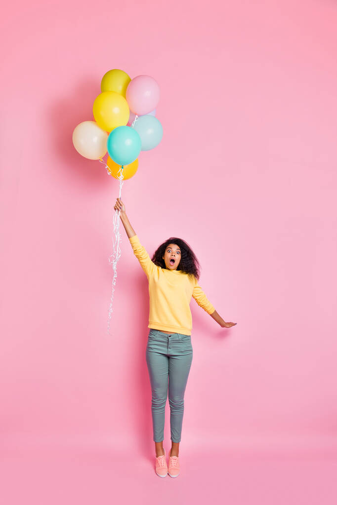 Foto vertical de cuerpo entero del hipster adolescente divertido emocional asombrado sosteniendo muchos globos en la mano sintiendo que puede ser volada hacia el cielo en las nubes en el día de verano aislado fondo de color pastel
 - Foto, imagen