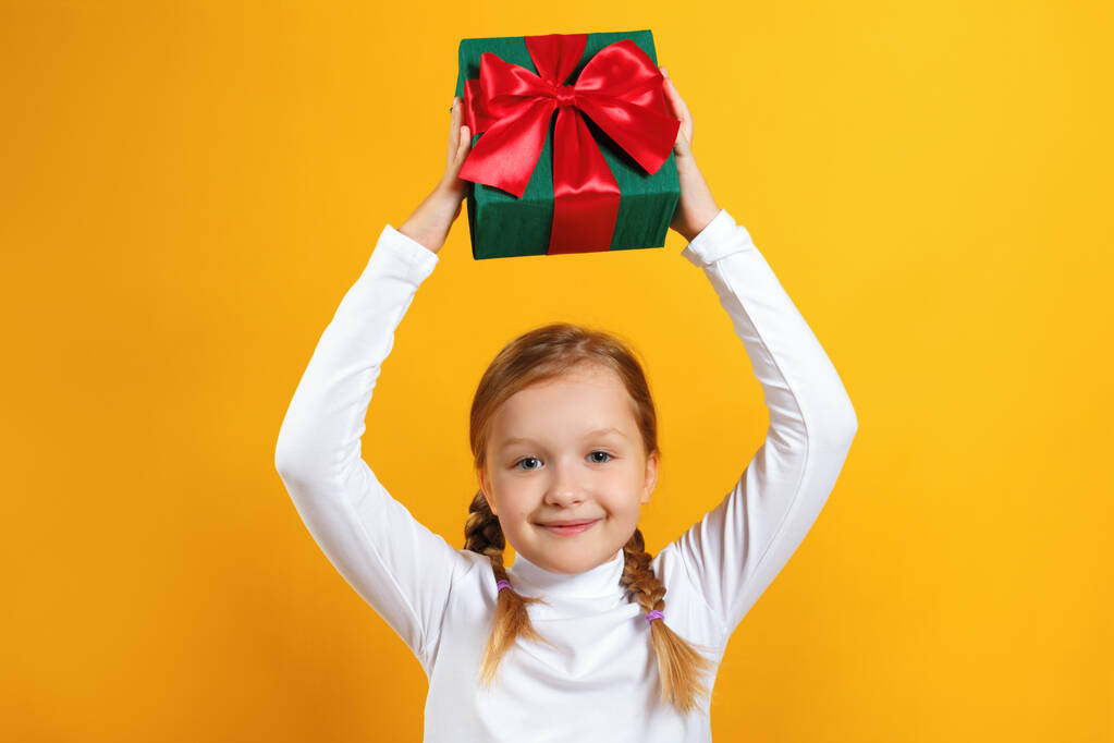 Schattig gelukkig meisje met een doos met een gave over haar hoofd. Een kind met staartjes in een witte coltrui op een gele achtergrond - Foto, afbeelding