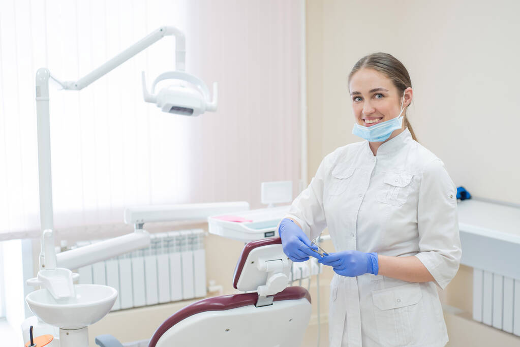 Médico feminino sorridente atraente em um uniforme branco no local de trabalho. Jovem dentista bonita em máscara e luvas está de pé no escritório perto da poltrona com ferramentas em suas mãos
 - Foto, Imagem