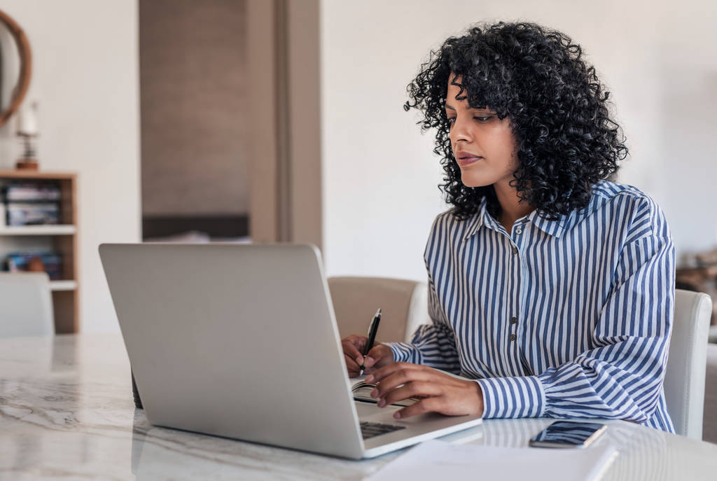 Молода жінка-підприємець використовує ноутбук і записує нотатки під час роботи з дому за обіднім столом
 - Фото, зображення