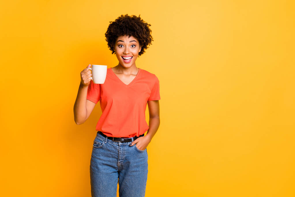 Sarı renkli arka planda izole edilmiş çay bardağı tutan turuncu t-shirt kot pantolon giyen sevimli, neşeli, çekici kız arkadaşın fotoğrafı. - Fotoğraf, Görsel