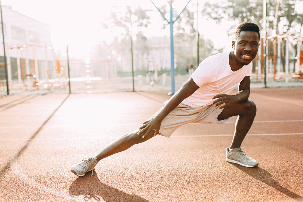 Атлетический афроамериканец делает упражнения на растяжку, готовится к утренней тренировке на стадионе
 - Фото, изображение
