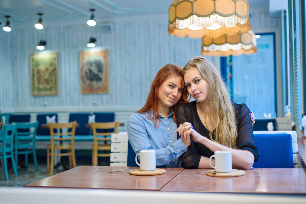 Однополые отношения. Счастливая лесбийская пара, сидящая в кафе. Девочки нежно держатся за руки и пьют кофе. Объятия любящих женщин. ЛГБТ. Два лучших друга в кафе
. - Фото, изображение