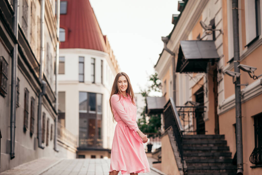 Ένα νεαρό, όμορφο κορίτσι με ένα ροζ φόρεμα περπατά στους όμορφους δρόμους της πόλης. Πολύ όμορφα πορτρέτα ενός κοριτσιού στην πόλη. - Φωτογραφία, εικόνα