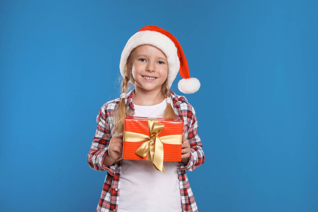 Happy little child in Santa hat with gift box on blue background. Christmas celebration - Photo, Image