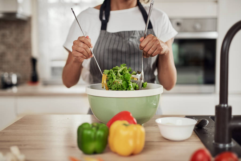 Atrakcyjne mieszane rasy kobieta w fartuchu mieszanie warzyw w misce stojąc w kuchni w domu. - Zdjęcie, obraz