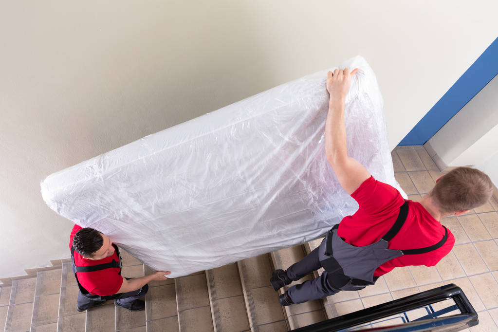Dois jovens homens se movimentam de uniforme levando o colchão embrulhado enquanto se movem para baixo da escada
 - Foto, Imagem