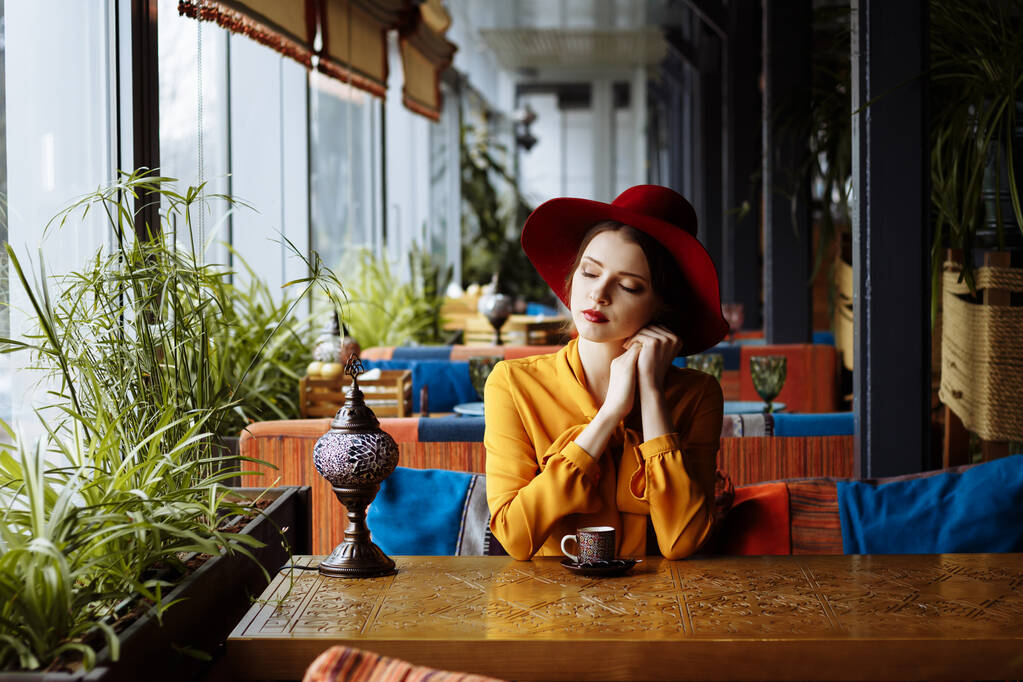 κορίτσι σε ένα καφέ με ένα φλιτζάνι καφέ και ένα καπέλ.πορτρέτο του αισθησιακό νεαρό κορίτσι φορώντας πλαδαρό καπέλο και μπλούζα με φιόγκο. Όμορφη μελαχρινή γυναίκα στο καφέ κρατώντας φλιτζάνι καφέ - Φωτογραφία, εικόνα