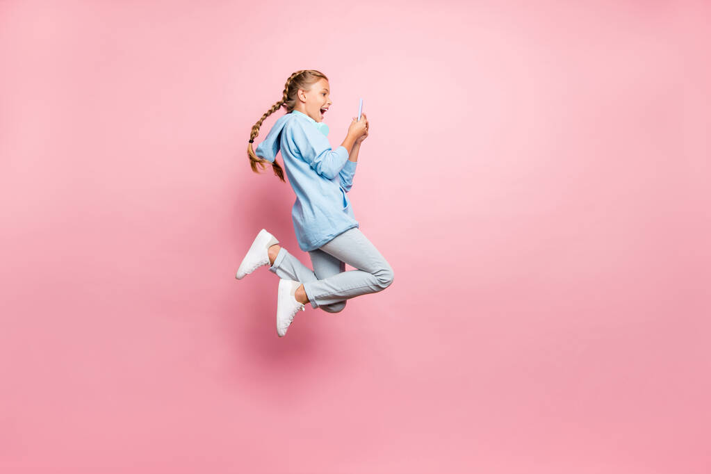 Ganzkörpergröße Foto von aufgeregt verrückt ekstatische Schulkind Browsing durch Telefon Springen tragen blaues Sweatshirt Pullover sportliche tragen Jeans Denim isoliert über rosa Pastellfarbe Hintergrund - Foto, Bild