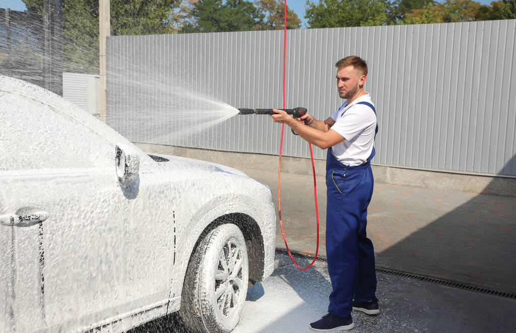 Молодий працівник прибирає автомобіль з струменем води високого тиску на автомийці
 - Фото, зображення
