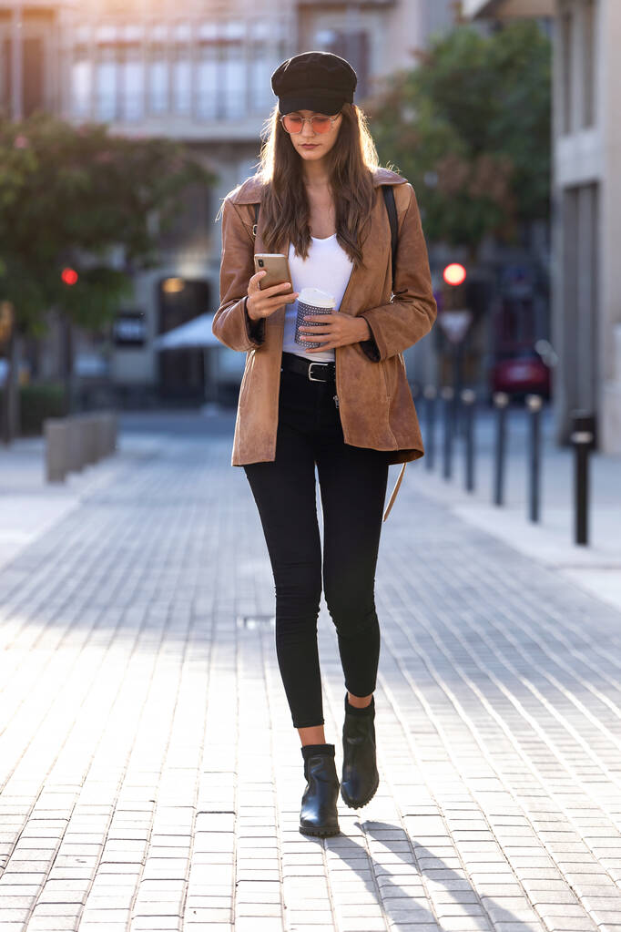 Ładna młoda kobieta używa telefonu komórkowego, trzymając kawę i stojąc na ulicy. - Zdjęcie, obraz