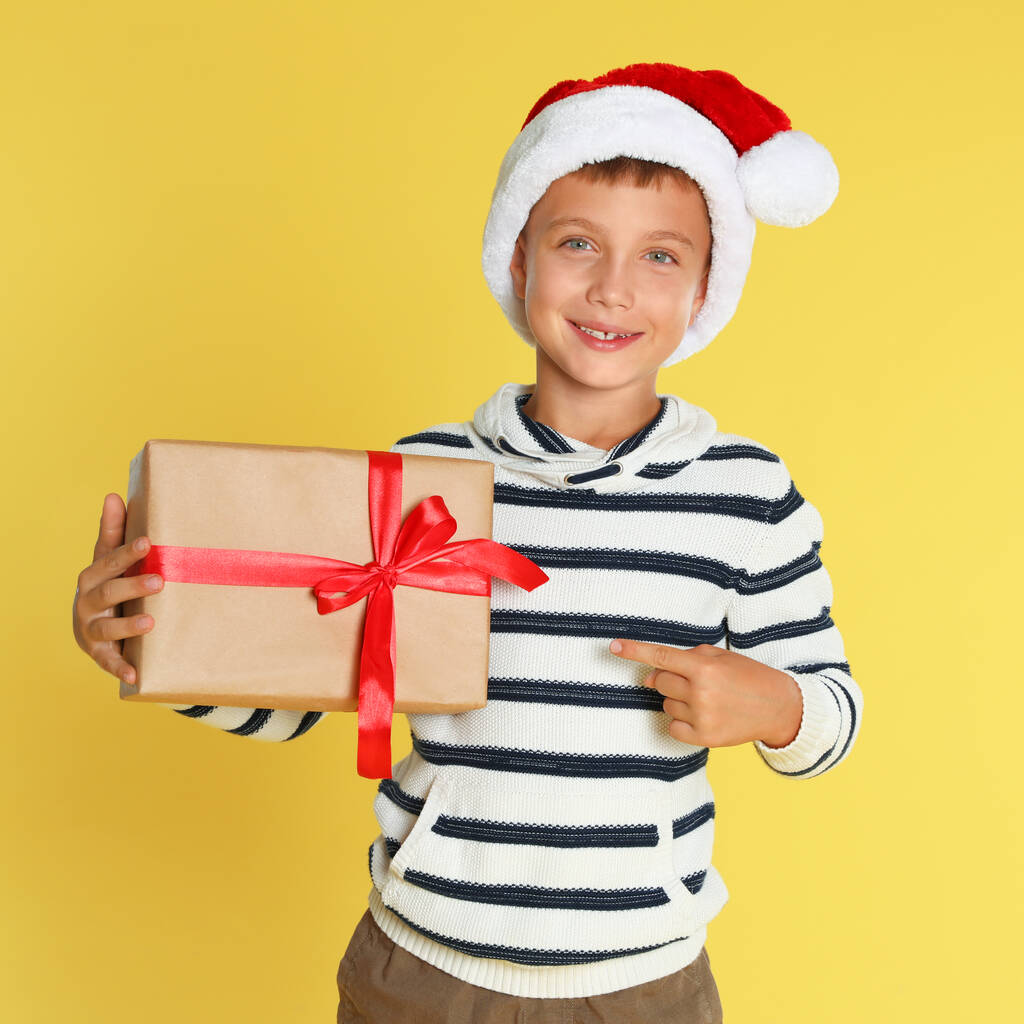 Cute little boy in Santa hat with Christmas gift on yellow background - Photo, Image