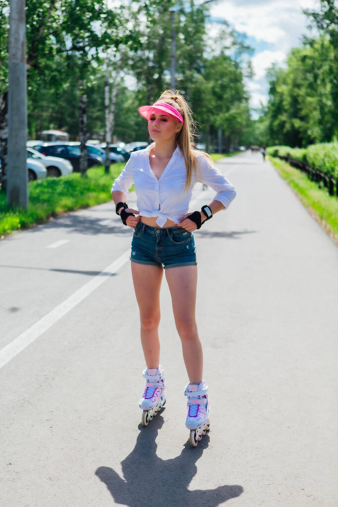 Retrato de uma menina emocional em uma viseira de boné rosa e luvas de proteção para patins e andar de skate em patins na estrada
. - Foto, Imagem