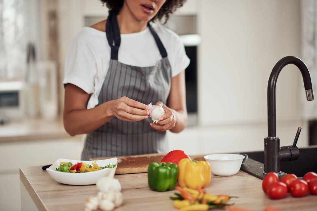 Nádherné smíšené závod žena v zástěře stojící v kuchyni a loupání česneku. - Fotografie, Obrázek