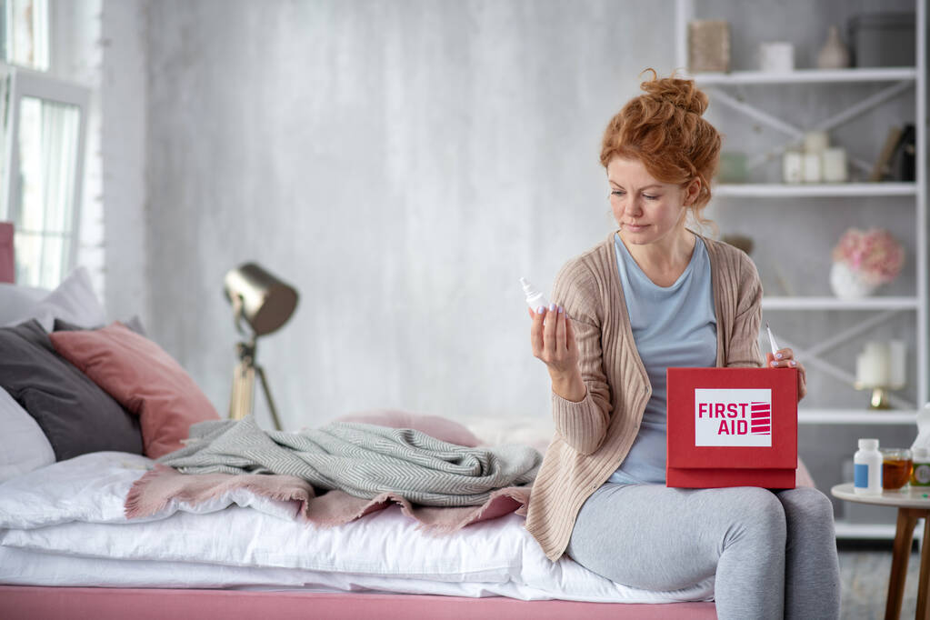 Mulher sentada na cama com kit de primeiros socorros e à procura de medicina
 - Foto, Imagem