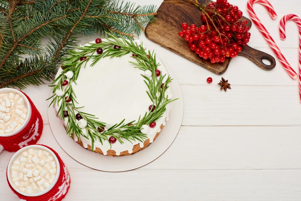 bovenaanzicht van kersttaart op witte houten tafel met snoepstokken, cacao, viburnum bessen en sparren takken - Foto, afbeelding