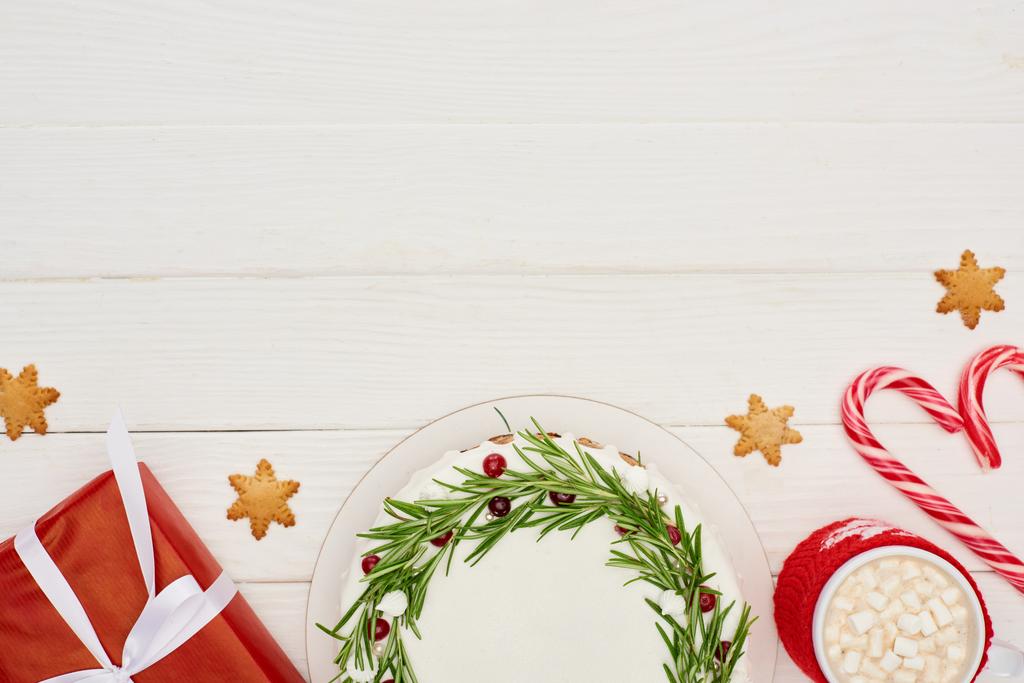 Draufsicht auf Weihnachtskuchen, rotes Geschenk, Zuckerstangen und Tasse Kakao mit Marshmallows auf weißem Holztisch - Foto, Bild