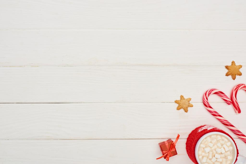 top view karácsonyi cukornád, kis ajándék, cookie-k és csésze kakaó mályvacukorral fehér fa asztalon  - Fotó, kép