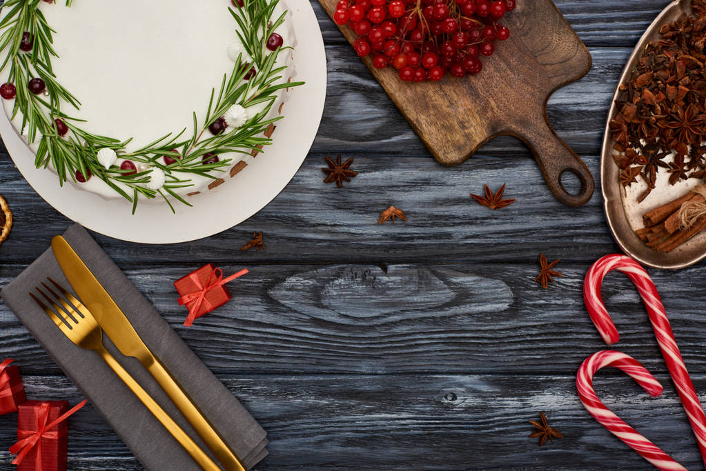 vrchní pohled na vánoční koláč s rozmarýnem a vibračními bobulemi, dárky, vidličkou a nožem na ubrousku na tmavém dřevěném stole  - Fotografie, Obrázek