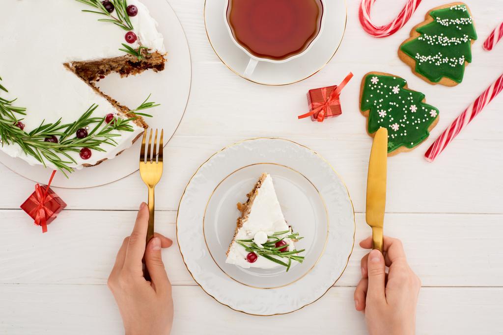 przycięty widok dziewczyny trzyma widelec i nóż do jedzenia ciasto świąteczne i ciasteczka z herbatą na białym drewnianym stole - Zdjęcie, obraz