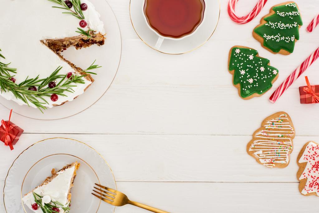 bovenaanzicht van kersttaart met rozemarijn, kopje thee en kerstboomkoekjes op witte houten tafel - Foto, afbeelding