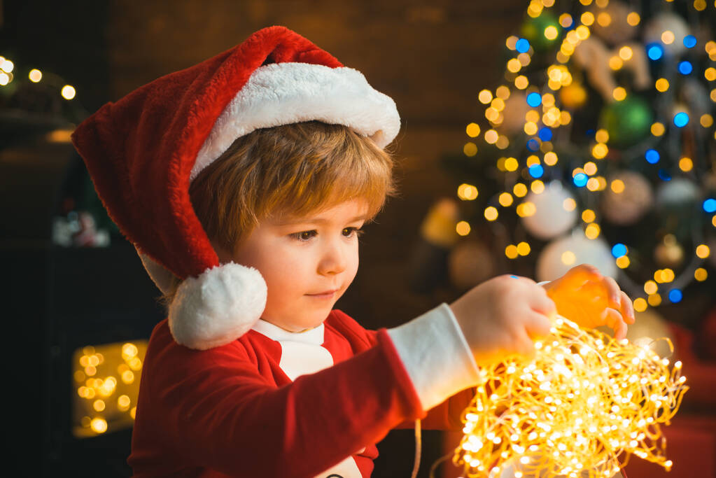 Kind mit Lichtergirlanden am Weihnachtsbaum und am Kamin an Heiligabend. Es ist ein Wunder. Familie mit Kindern feiert Weihnachten zu Hause. Junge niedliches Kind fröhliche Stimmung spielen in der Nähe von Weihnachtsbaum. - Foto, Bild